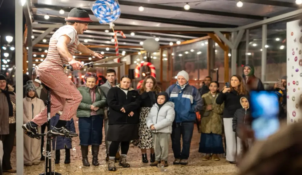 A street performer dressed in a striped shirt, red suspenders, and striped trousers balances on a unicycle while juggling metal pans at a festive Hanukkah event on the Armon HaNatziv Promenade in Jerusalem.